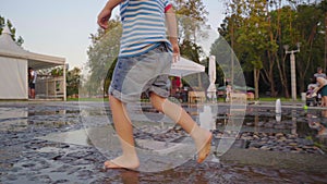 A little boy in denim shorts and yellow shoes runs in the fountain on summer evening