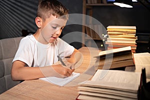 Little boy doing homework at home late in evening, schoolboy writes a test with a pen in a notebook while sitting at a