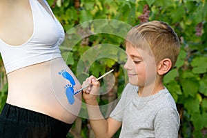 Little boy draws with a brush on the belly of his pregnant mom babyâ€™s footprint. Pregnant concept. Close-up