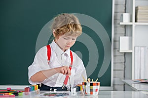 Little boy draws in classroom sitting at a table, having fun on school blackboard background. Kids early arts and crafts