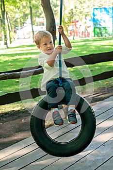 A little boy dressed in a white T-shirt and jeans is riding a swing made of a car tire in a summer park