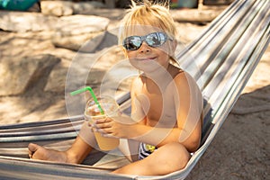 Little boy, drinking coctail, orange juice on the beack in hammock, enjoying summer