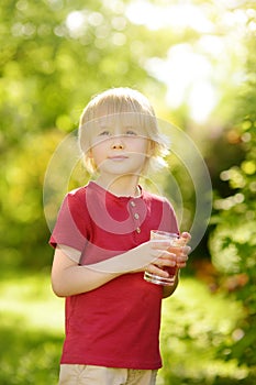 Little boy drinking glass of water on hot sunny summer day in the backyard or home garden. Feeling of thirst
