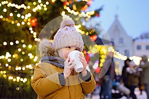 Little boy is drinking hot chocolate or child mulled wine near giants Christmas fir tree