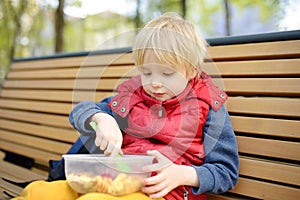 Little boy is eating his lunch after kindergarten or school from plastic container on bench in the park. Street take away food for