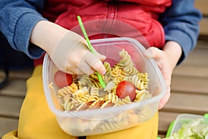 Little boy is eating his lunch after kindergarten or school from plastic container on bench in the park. Street take away food for