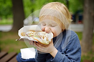 Little boy eating hot dog in public park. Child enjoying his to go meal outside. Fast food is a junk food. Overweight kids