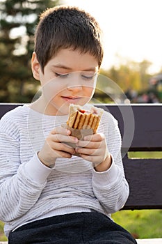Little boy eating a hotdog while sitting on a bench in the park. Street fast food.