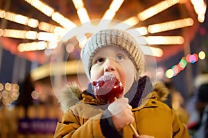 Little boy eating red apple covered in caramel on Christmas market. Traditional child& x27;s enjoyment and fun during Xmas time