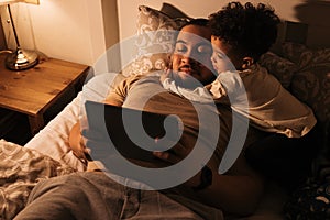 Little boy embracing his father while them lying on a bed. Young father reading book to his child from a digital tablet before