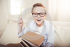 Little boy in eyeglasses holding pile of books and pointing up with pencil