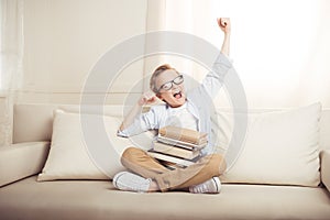 Little boy in eyeglasses sitting on sofa with books stretching and yawning