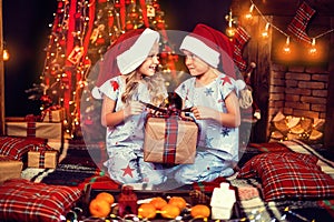 Little boy and girl in pajamas opening gift box while sitting near fireplace and Christmas tree