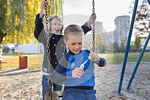 Little boy and girl playing in autumn park, children sitting on swing blow soap bubbles