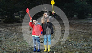 A little boy and girl in rubber boots are walking through the forest and holding shovels.