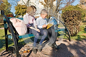 Little boy and girl schoolchildren reading book, sitting on bench, children with backpacks