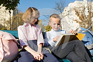 Little boy and girl schoolchildren reading book, sitting on bench, children with backpacks