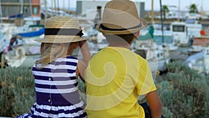 Little boy and girl are sitting and looking on the quay of the yacht port