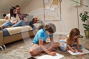 Little boy and girl sitting on the wooden floor and drawing with colorful pencils while their parents, happy mother and