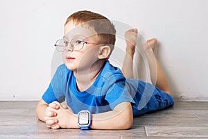 A little boy in glasses and in a blue t-shirt with a blue smart watch on his hand is lying on the floor on a white