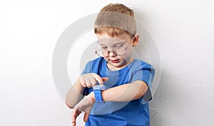 A little boy with glasses and a blue T-shirt looks at his smart watch against the background of a white wall.