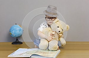 A little boy in glasses and a hat sits on the floor and hugs a teddy bear.