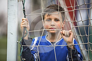 Little boy in goalkeeper uniform behind mesh football goal on the stadium.