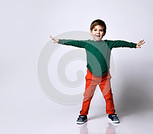 Little boy in green jumper, orange pants, blue sneakers. He raised hands and looking overjoyed, posing  on white