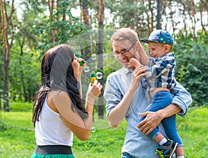 A little boy in the hands of the dad and and his mom blowing soap bubbles.