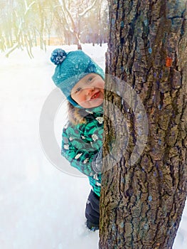 A little boy happily smiles peeking from behind a tree trunk on a winter day