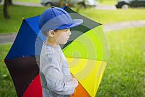 Little boy in hat with multicolor umbrella in park after rain on sunny summer day