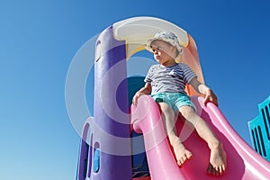 A little boy in a hat is ready to slide, on a colourful slide, he looks into the distance. There is free space for text in the