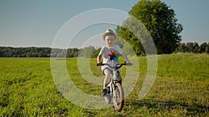 Little boy in hat is riding a bike with windmill on green field in country road.