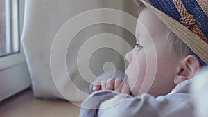 little boy in hat sits at window, looks sadly out window, watches life outside