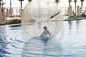 Little boy have fun inside big plastic balloon on the water of swimming pool on the summer resort. Kid inside big inflatable