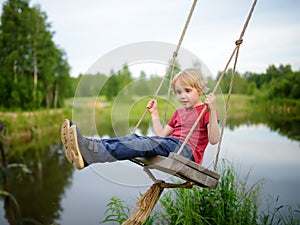 Little boy having fun on a swing hanging on big tree near pond or river in the forest in summer day