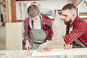 Little boy helps his carpenter father measuring the wooden plank in workshop.