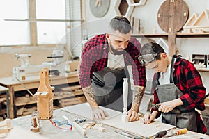 Little boy helps his carpenter father measuring the wooden plank in workshop.