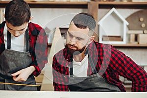 Little boy helps his carpenter father measuring the wooden plank in workshop.