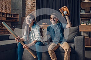 Little boy and his beautiful mother sitting on couch in living room with baseball bat and mitt while