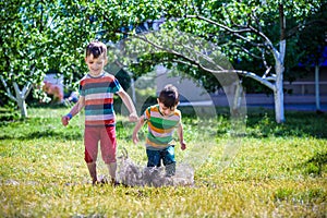 Little boy and his brother play in summer park. Children with colorful clothes jump in puddle and mud in the garden.