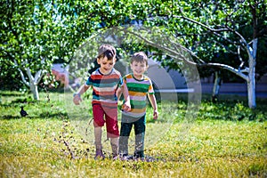 Little boy and his brother play in summer park. Children with colorful clothes jump in puddle and mud in the garden.