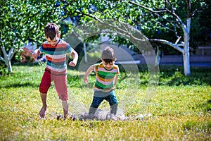 Little boy and his brother play in summer park. Children with colorful clothes jump in puddle and mud in the garden