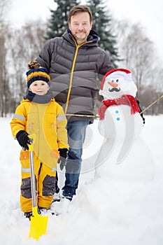 Little boy with his father building snowman in snowy park. Active outdoors leisure with children in winter