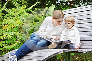 Little boy and his mother sitting on bench in park and reading