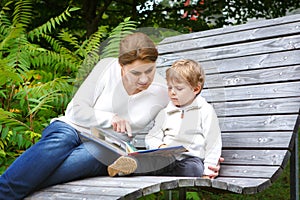 Little boy and his mother sitting on bench in park and reading b