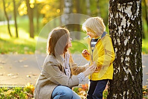 Little boy with his young mother having fun during stroll in the public park at sunny autumn day. Active family time on nature.