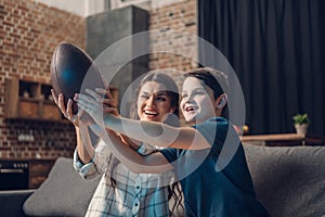 Little boy and his young mother holding a rugby ball while watching tv on couch