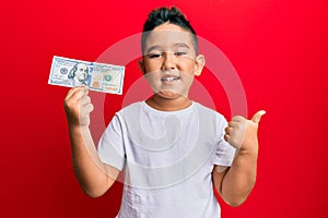 Little boy hispanic kid holding 100 dollars banknote smiling happy and positive, thumb up doing excellent and approval sign