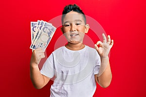 Little boy hispanic kid holding 5000 japanese yen banknotes doing ok sign with fingers, smiling friendly gesturing excellent